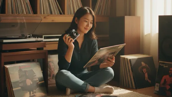 20대 한국 여성의 아날로그 라이프스타일. 따뜻한 조명 아래 카펫 위에 앉아 필름 카메라(35mm)를 든 채 바이닐 레코드(LP) 커버를 감상하는 모습. 빈티지 스피커와 턴테이블이 보이는 방 안에 수백 장의 LP가 가득하며, 차분한 색감과 얕은 심도로 향수를 자극하는 포토 리얼리즘 화보.